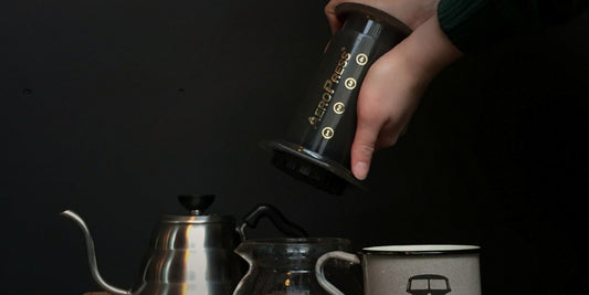 A hand holding an AeroPress beside a gooseneck kettle and mug in dramatic light