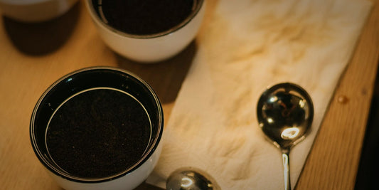 Cupping bowls of ground coffee and a cupping spoon on linen, lit warmly against a dark background
