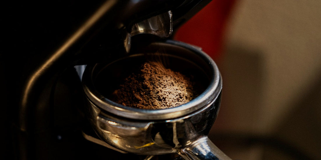 A burr grinder dosing fresh grounds into a portafilter, lit low against a dark background