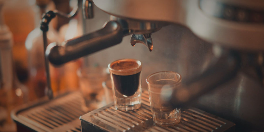Two glasses filling with espresso under a machine, lit warmly against a dark background