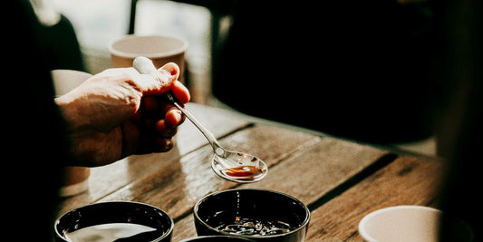 A hand holding a cupping spoon over cups of coffee on a dark wooden table