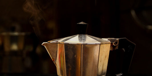 A moka pot steaming on a stove, lit warmly against a dark background