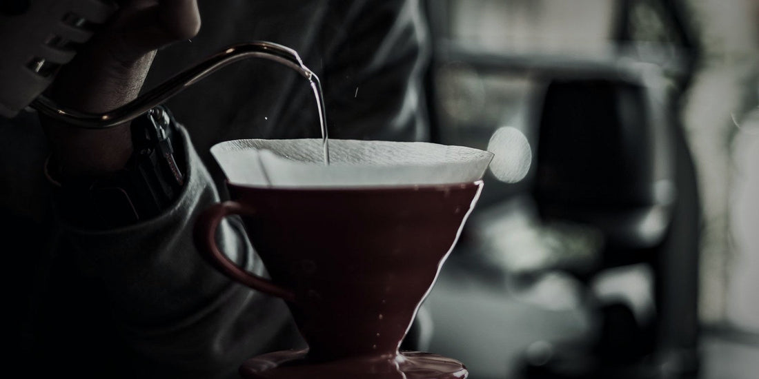 A gooseneck kettle pouring into a cone dripper, lit dramatically against a dark background
