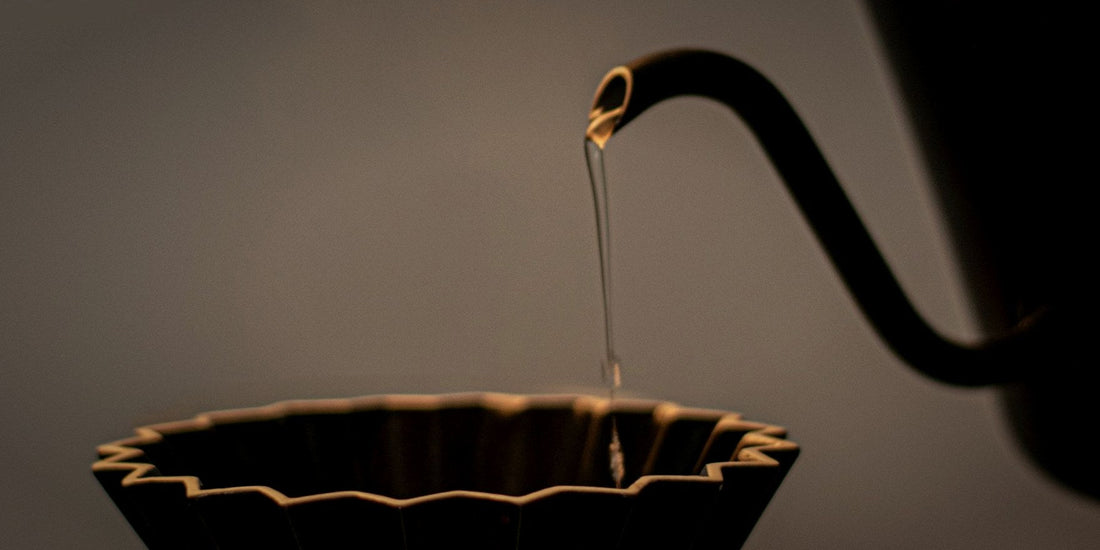 A gooseneck kettle pouring water into a dark coffee dripper, lit dramatically